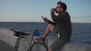 Man cyclist resting overlooking sea, drinking coffee and using smartphone while sitting on embankment. Male 30s wearing sunglasses, drinking takeaway coffee and using mobile phone on waterfront. video