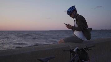 Theme is cycling in seaside. Male cyclist stopped to enjoy the sea view and see the road on the map on his smartphone. A male with a bicycle on the embankment is resting and using a smart phone. video