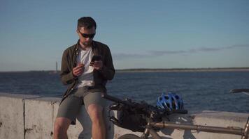 Bicyclist having meal and browsing websites on smartphone while ride bike near lake. Man eating lunch and using phone in front of pond. Outdoors on his bicycle for lunch. Snacking while cycling. video