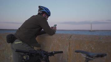 Theme is cycling in seaside. Male cyclist stopped to enjoy the sea view and see the road on the map on his smartphone. A male with a bicycle on the embankment is resting and using a smart phone. video
