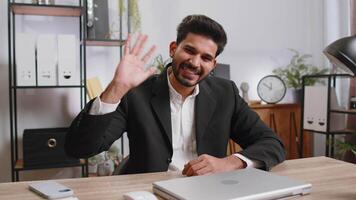 Businessman working on laptop smiling friendly at camera and waving hands gesturing hello at office video