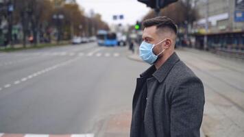 Man in mask on city transport background. Side view of a man in warm coat and protective medical mask standing near the road. Slow motion. Virus protection. video