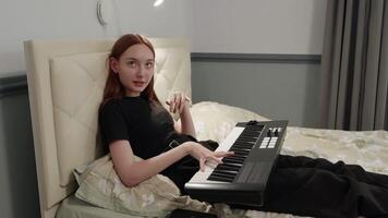 Teenage girl enjoying a creative moment while composing music on a midi keyboard, comfortably seated on her bed in a cozy bedroom filled with warm evening light video