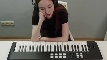 Bored young female composer with headphones is sitting at a white table, resting her head on her hand and looking for inspiration at a midi keyboard video