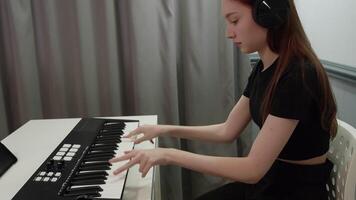 Young female musician with headphones playing a midi keyboard while composing music, sitting at a white table in her home studio, using a tablet as support video