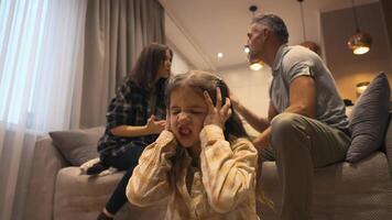 Desperate american 7-8 year old girl during parents quarrel closing ears at home. A little girl is sitting on the floor and covering ears with hands while mother and father arguing on background video