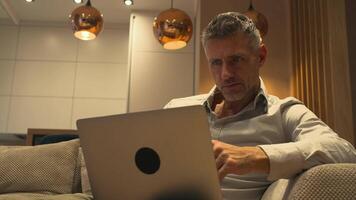 A gray-haired handsome man is sitting with his laptop at home in business outfit and working, thinking and trying to make sense of crypto video