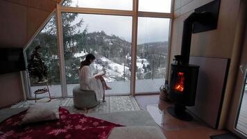 Woman relaxing by a fireplace with a snowy forest view from a cozy room, wrapped in a robe. video
