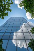 Looking up at a sleek glass building, vibrant green leaves frame the view, while fluffy white clouds reflect off the shiny surface beneath a clear blue sky. photo