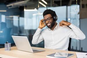 Young man at workplace working with laptop, listening to music, smiling and singing along. Worker in headphones happily resting and relaxing while working in the evening. photo