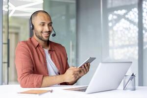 Man with headset working inside office, using tablet computer and laptop. Support service worker at workplace, consulting and helping clients, online remotely. photo