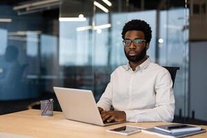 Portrait of a young serious man inside the office with a laptop. Businessman looking at the camera, working in the evening at the workplace. photo