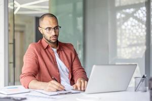 A professional sitting at a desk working with a laptop and writing notes, conveying concentration and productivity in a bright office environment. photo