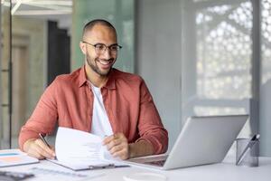Successful financier doing paperwork inside office. Smiling man working with contracts invoices and reports. Using laptop to fill out tax forms. photo