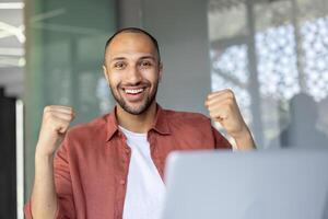 A cheerful individual in an office environment expressing joy with raised fists, representing success and positivity. The light-filled background complements sense of accomplishment conveyed. photo