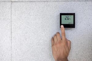 A person adjusts a digital thermostat mounted on a textured wall, displaying degrees Celsius. photo
