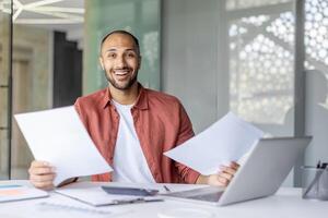 Portrait joyful successful financier, man with documents and financial reports in his hands smiles and looks at the camera. Businessman works inside the office with bills, satisfied with the results. photo