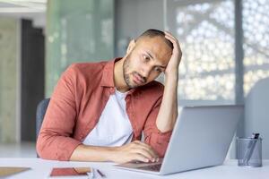 A man sitting at a desk in a modern office, appearing frustrated while using his laptop. The scene conveys stress, problem-solving, and difficulty in a work-related context. photo