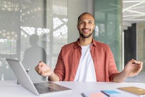 A young man in a casual shirt smiling and meditating at a desk with a laptop in a bright and modern office environment. The mood conveys calmness and balance during work hours. photo