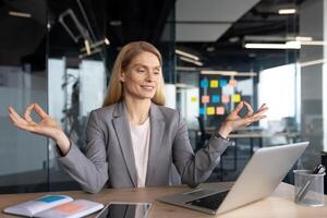 Joyful woman meditating in lotus pose inside office at workplace. Mature businesswoman in business suit, resting and relaxing watching online yoga course, using laptop. photo