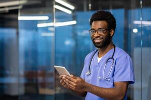 Portrait of a successful doctor with a tablet computer in hands, man smiling and looking at camera. The doctor is inside office of modern clinic, working in evening, consulting patients remotely. photo