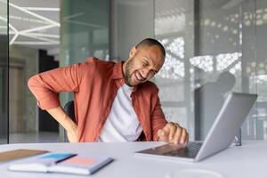 Man experiencing back pain while working at office desk with laptop. Concept of discomfort, posture issues, and need for ergonomic workspace during work. Focus on health. photo