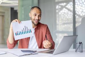Smiling professional explaining statistical data while holding a chart and using a laptop, showcasing modern business communication. photo
