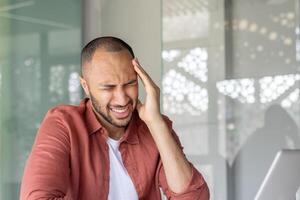 A man sitting indoors with a pained expression, holding his head as if experiencing a headache or stress, dressed casually and with a modern context suggesting work or personal challenge. photo