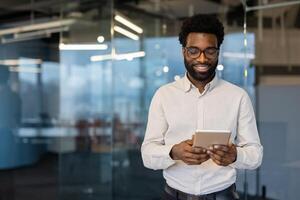 Successful and smiling man standing with tablet computer near window, businessman browsing social networks, educational materials. Office worker at workplace inside office. photo