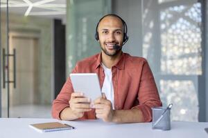 A man with a headset and a tablet computer in his hands looks into the camera, a support service worker at work, advising clients. Web camera view. photo