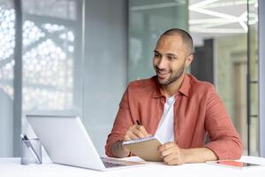 Man watching online webinar, business conference remote meeting. Employee studying sitting at desk inside office, writing data in notebook, smiling satisfied. photo