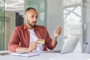 Upset and deceived man at workplace holding bank credit card in hands, received error of funds transfer blocking account, fraudulent actions. Working inside office with laptop. photo