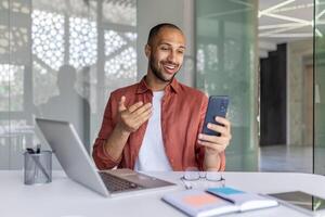 A joyful man sits inside the office with a phone in his hands, uses an application for online remote communication. Office worker with a laptop at the workplace. photo