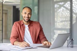 Portrait of a young successful businessman financier at his workplace inside the office, the employee is smiling and looking at the camera, writing down data on a paper report document photo