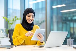 Successful woman in hijab using app on tablet computer. Arab office worker smiling contentedly, working inside office at workplace. photo