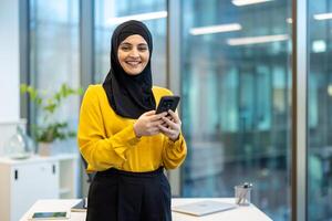 Portrait of a successful office worker inside office, a woman in hijab smiling and looking at camera. Businesswoman holding a phone in her hands, typing a message and using an online application. photo