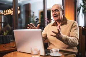 Senior man talking and gesturing while having a call on his laptop. He is using computer and drinking coffee and relaxing in the city cafe. photo