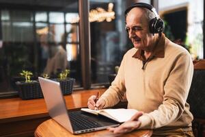 Senior man using wireless headphones, laptop and taking notes while attending online class. He is talking and gesturing during a call on computer in the cafe. photo