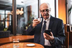 Senior businessman in formal wear using smart phone while enjoying drinking coffee in the bar. photo