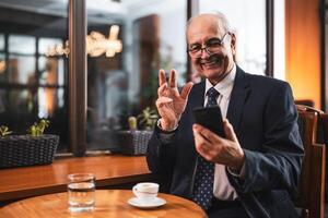 Senior businessman smiling and gesturing while holding mobile phone and sitting at the table in cafeteria. photo