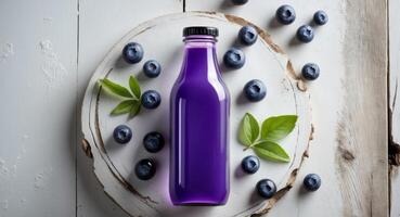 Vibrant violet blueberry juice bottle centered on white wooden board surrounded by fresh blueberries and green leaves viewed from above. photo