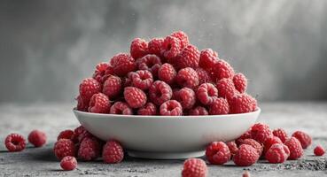 Fresh red raspberries overflowing in a white bowl on a textured gray concrete background with scattered berries and ample copy space. photo