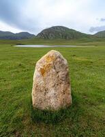 Single standing stone in a grassy field with mountains in the background photo