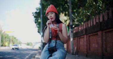 Handheld low angle view shot, Happy young Asian woman sits drinking iced tea next to a brick wall, smiling and enjoying the atmosphere around. video
