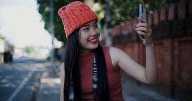 A young woman wearing an orange beanie poses for a selfie and smiles happily while sitting outdoors on a sidewalk. video