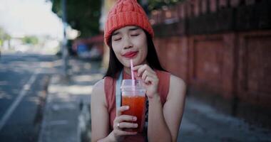 Handheld selective focus shot, Happy young Asian woman sits drinking iced tea next to a brick wall, smiling and enjoying the atmosphere around. video