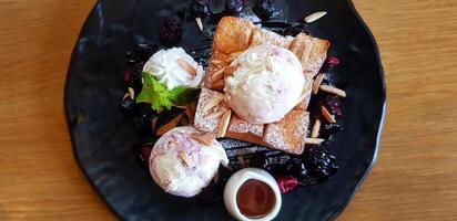Top view or flat lay of yogurt ice cream with toast bread, sliced almond, whipped cream, small white honey mug, berry sauce and mint leaves on top in black plate. Refreshment and sweet dessert concept photo