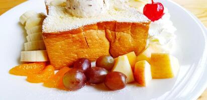 Close up sweet butter toast with top of chocolate chip ice cream sprinkle icing sugar, whipped cream with red cherry on top, vanilla ice cream and fresh fruit in white plate on wooden table. photo