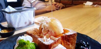 Toast Butter Bread, vanilla ice cream and sliced almond or nut pour with honey sauce on black dish. Close up Sweet dessert on wooden table at cafe with copy space and blurred background. photo