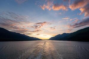 Puffs of clouds painted with colors of pink, orange, and purple, floating peacefully over a body of water at sunset with mountains in the background. photo